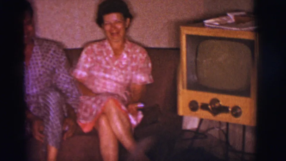 A 1966 home interior frame shows the woman smiling on the couch, the man partly cropped, and a wood-cabinet television beside them.