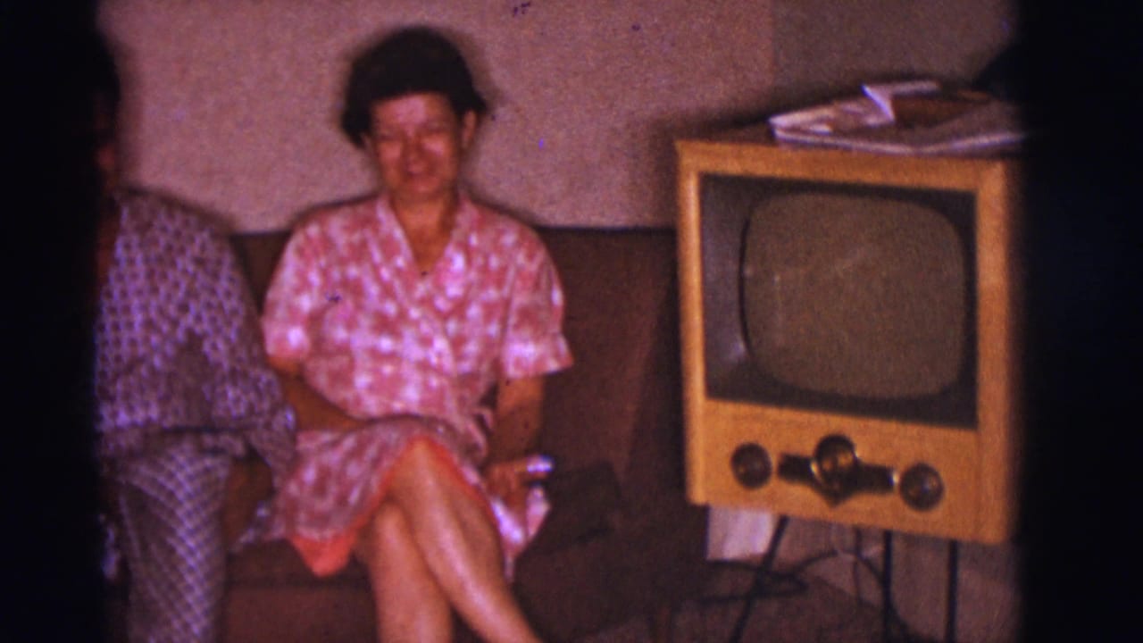 Woman in a pink patterned dress sits on a couch beside a wooden television set.