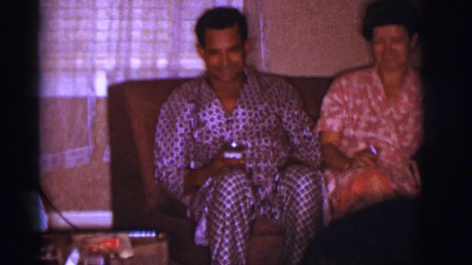 Two adults in patterned clothing sit together on a couch in a 1966 Clarkdale living room, with the dog mostly out of frame in front.