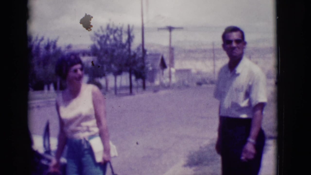 Woman with a handbag and man in sunglasses stand on a sunny street near a car.