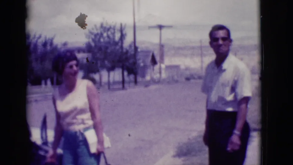 In 1966 Clarkdale, Arizona, a woman with a handbag and a man in sunglasses stand beside a car on a bright street.