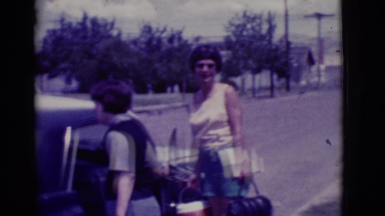 Two women stand beside an open car trunk with bags on a sunny street.