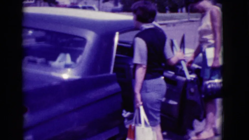 A woman in a vest and skirt holds a white bag beside the rear of a classic car during a 1966 Clarkdale luggage-loading moment.