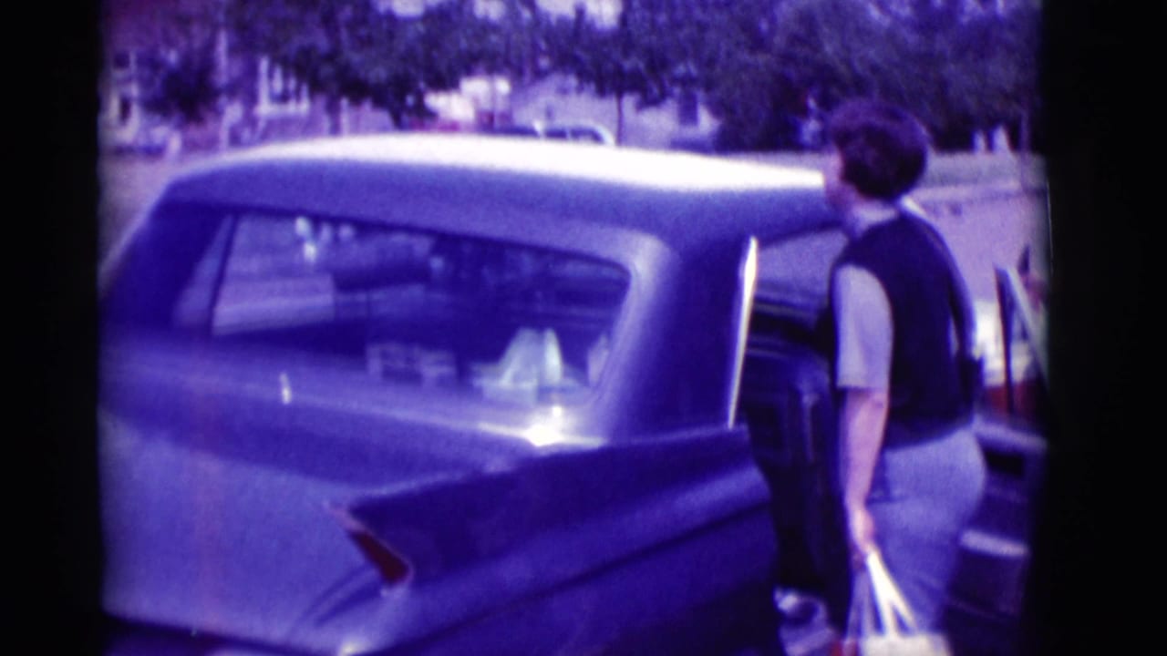 Woman with shopping bag stands beside a dark finned sedan on a sunny street.