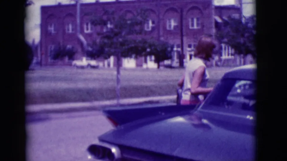 In 1966 Clarkdale, Arizona, a woman stands beside the dark sedan with trees, lawn, and a brick building across the street.