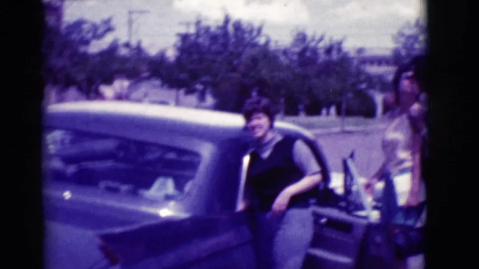 In 1966 Clarkdale, Arizona, two women stand by the open door of a dark classic sedan on a sunny street.