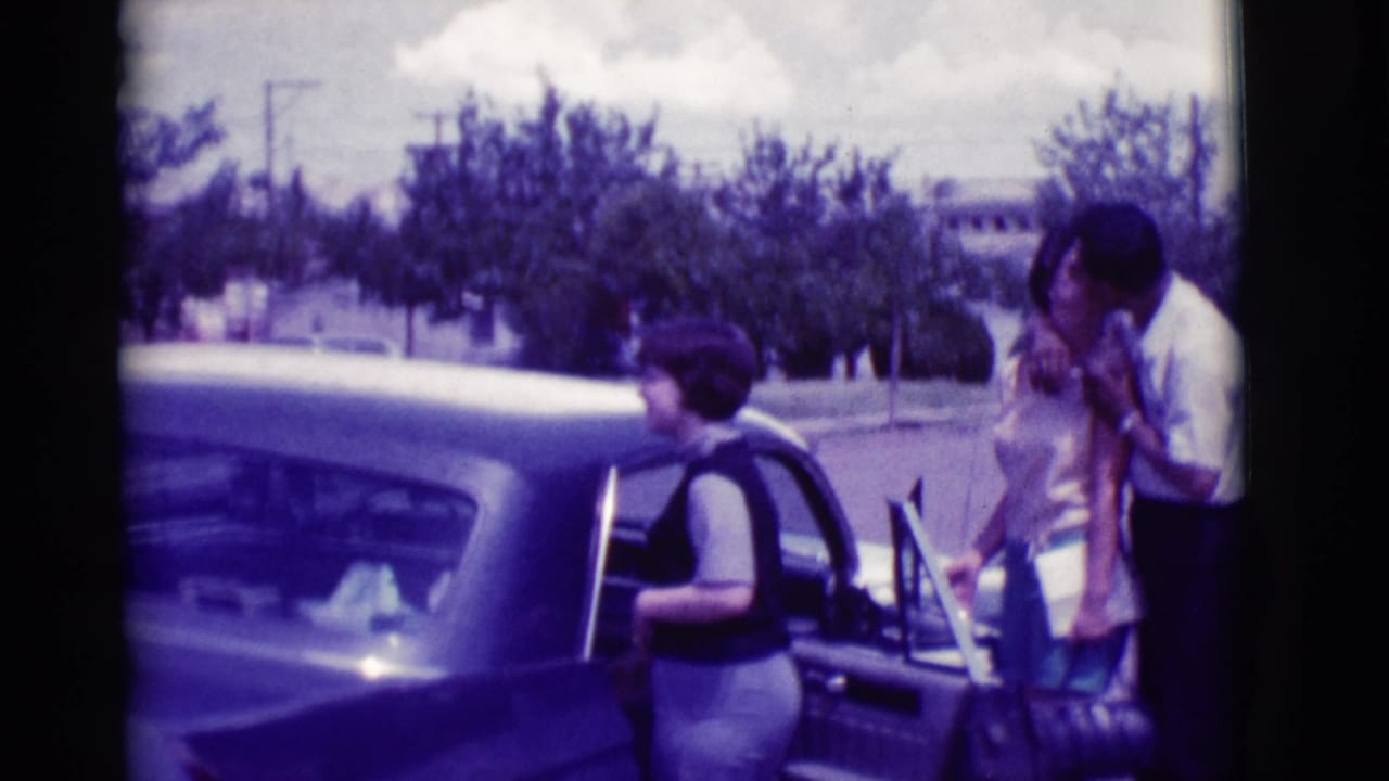 Adults beside an open classic sedan door on a suburban street.