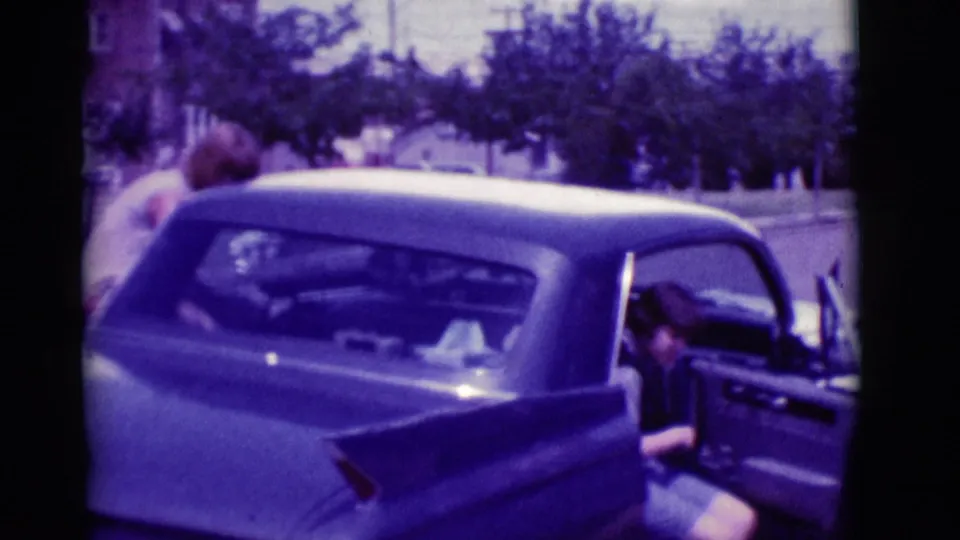 The rear of the classic sedan fills the frame as a woman sits or settles into the back seat with the door still open.