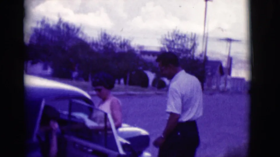 A woman stands beside the open rear door of the classic sedan while a man in a white shirt waits on the street in 1966 Clarkdale footage.