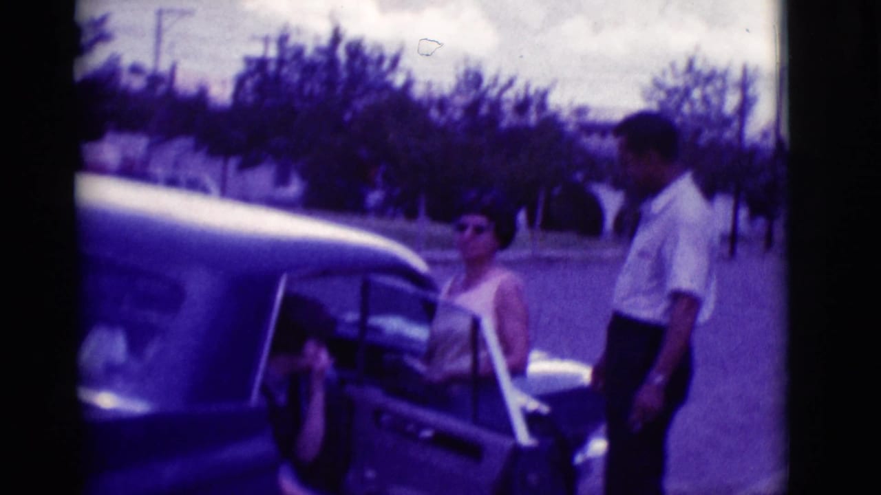 Adults beside a classic car with an open door on a tree-lined street.