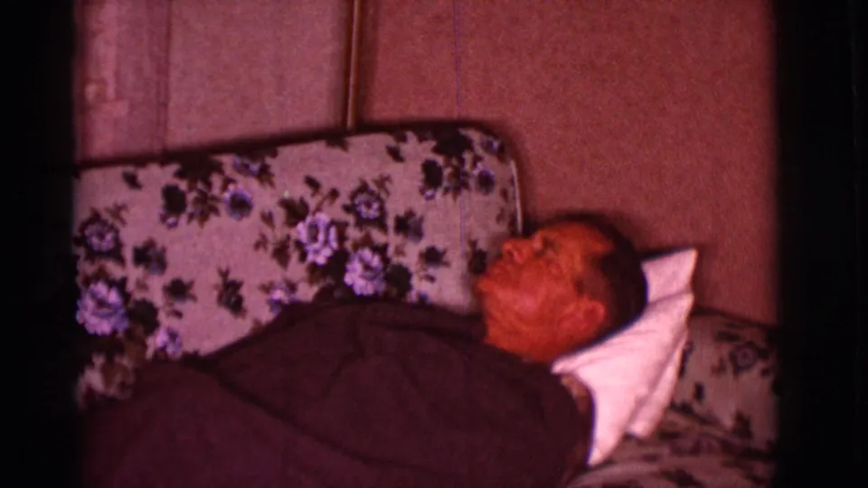 The 1966 home-movie frame shows the man reclined on the floral-patterned sofa with a pillow behind his head.