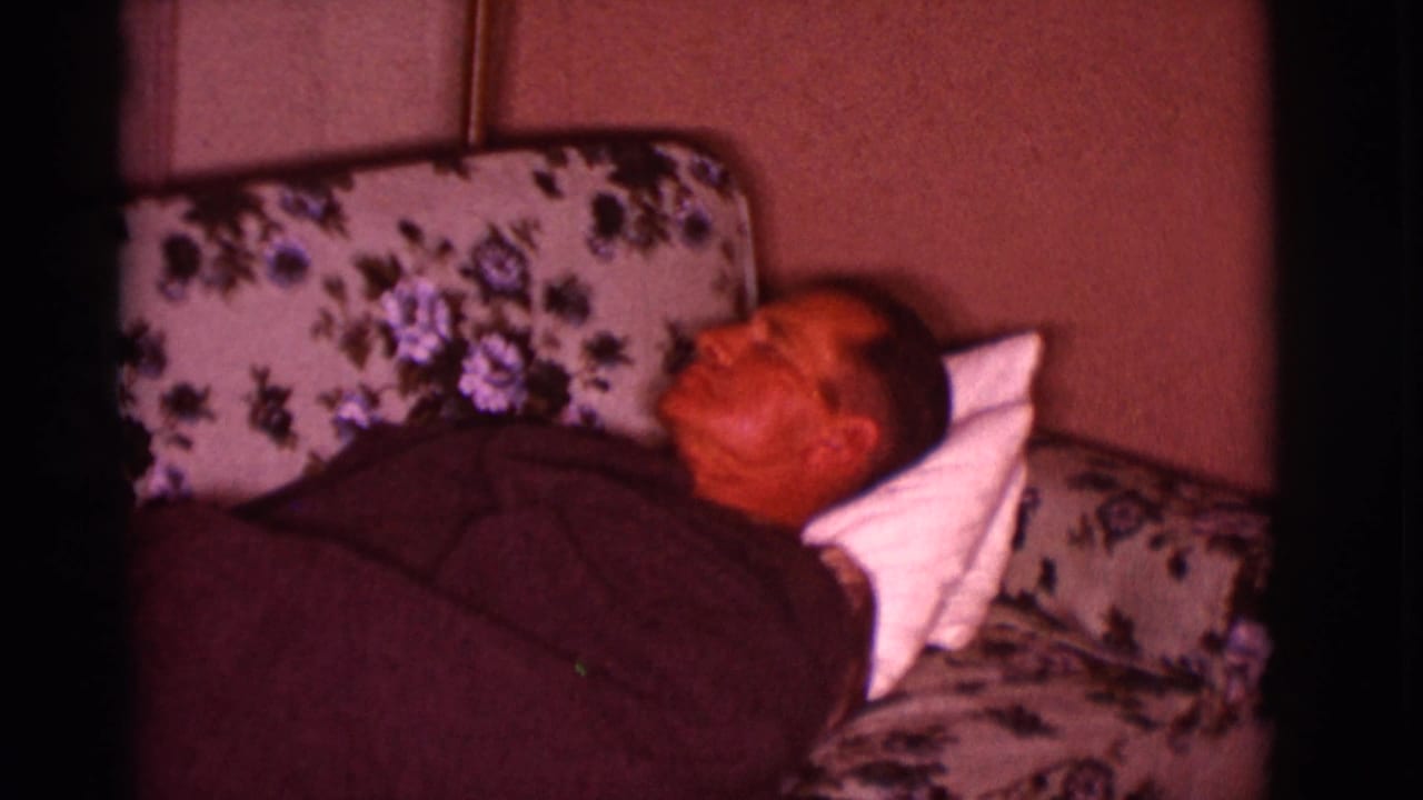 Adult man sleeping on a floral couch under a dark blanket with a white pillow.