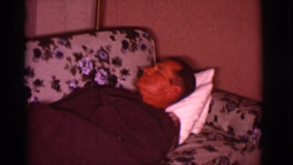 In a 1966 Clarkdale, Arizona home interior, an adult man sleeps on a floral couch beneath a dark blanket.