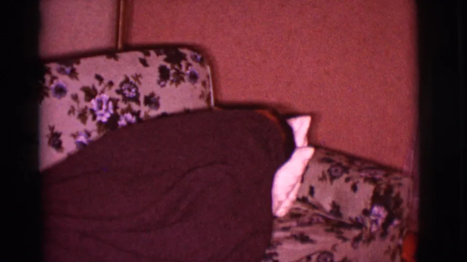 A blanket-covered adult lies turned away on a floral couch, with only bedding and the domestic interior clearly visible.
