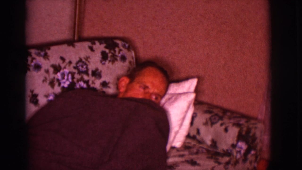 Man lying under a dark blanket on a floral couch with a white pillow.