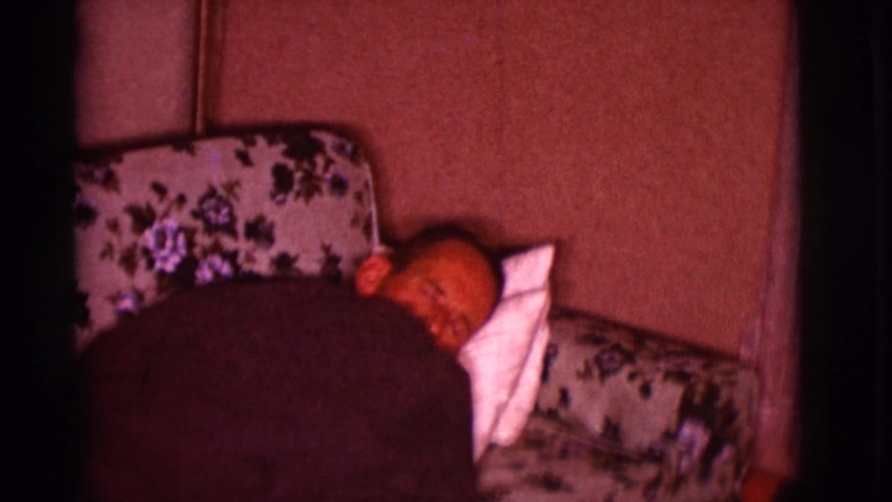 Man resting on a floral couch, partly covered by a dark blanket.