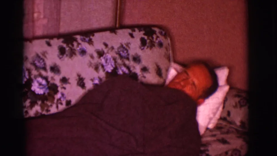 The man is shown farther along the couch, still covered by a dark blanket in the dim 1966 Clarkdale home interior.