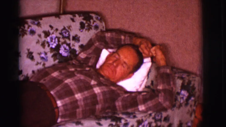 The frame shows the man reclining on the floral couch, eyes closed or lowered, with a white pillow under his head and plaid shirt visible.