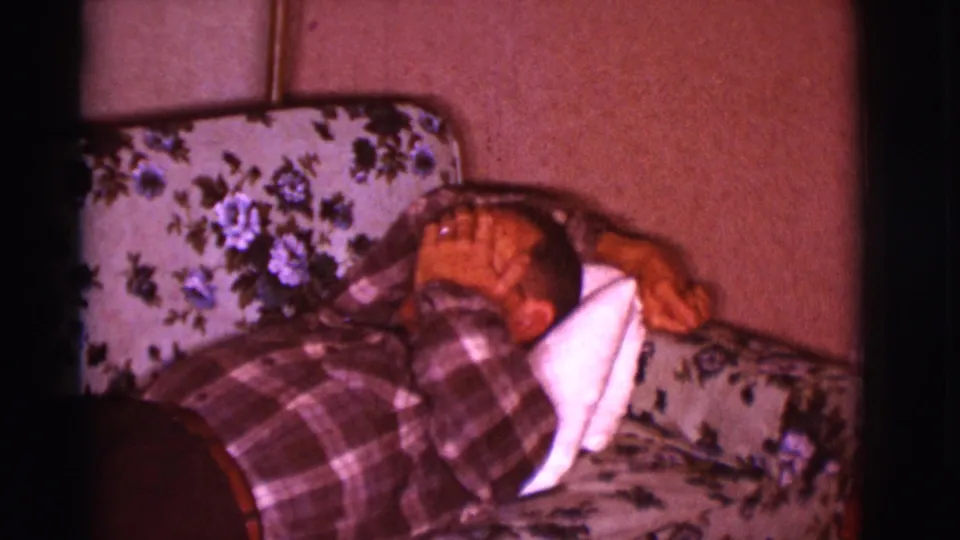 The man lies on the floral couch with a white pillow under his head and one hand raised across part of his face in a private interior.