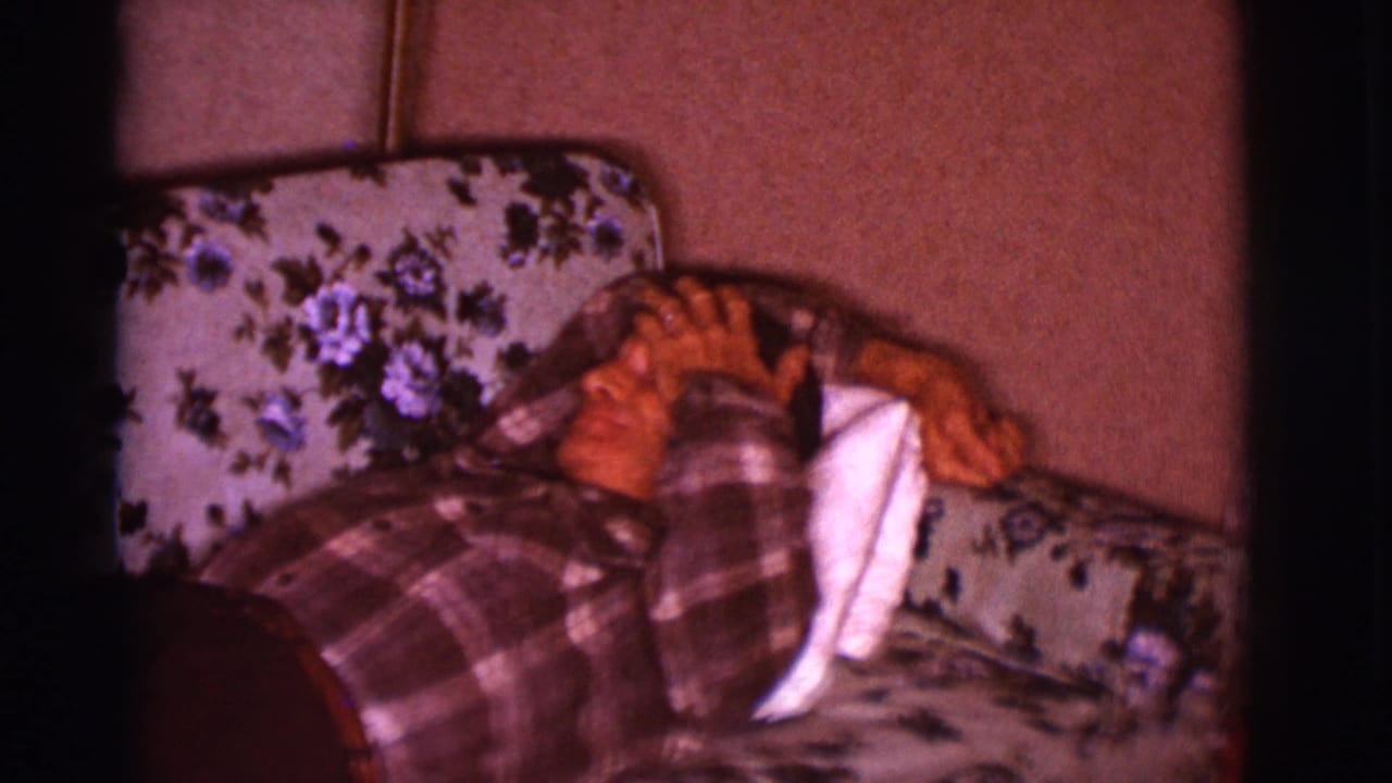 Older man in a plaid shirt lying on a floral couch with his hand near his face.