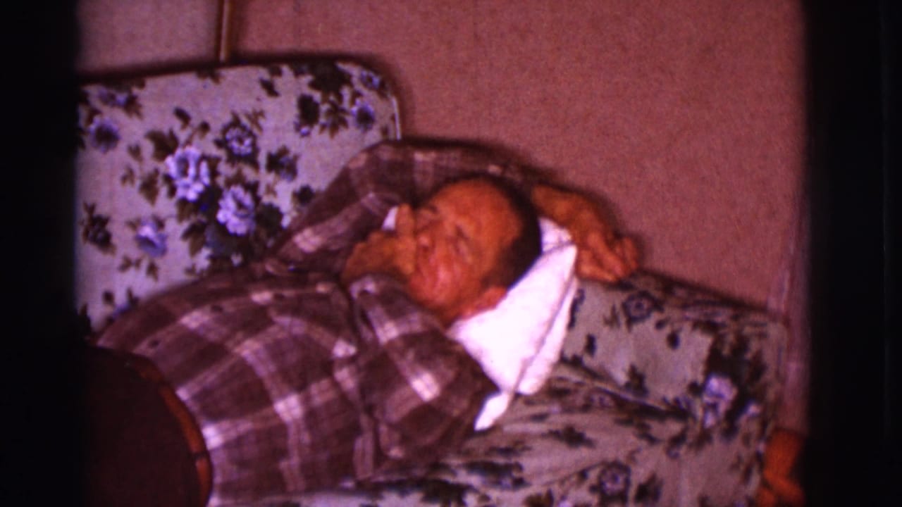 Older man reclining on a floral couch with a white pillow behind his head.