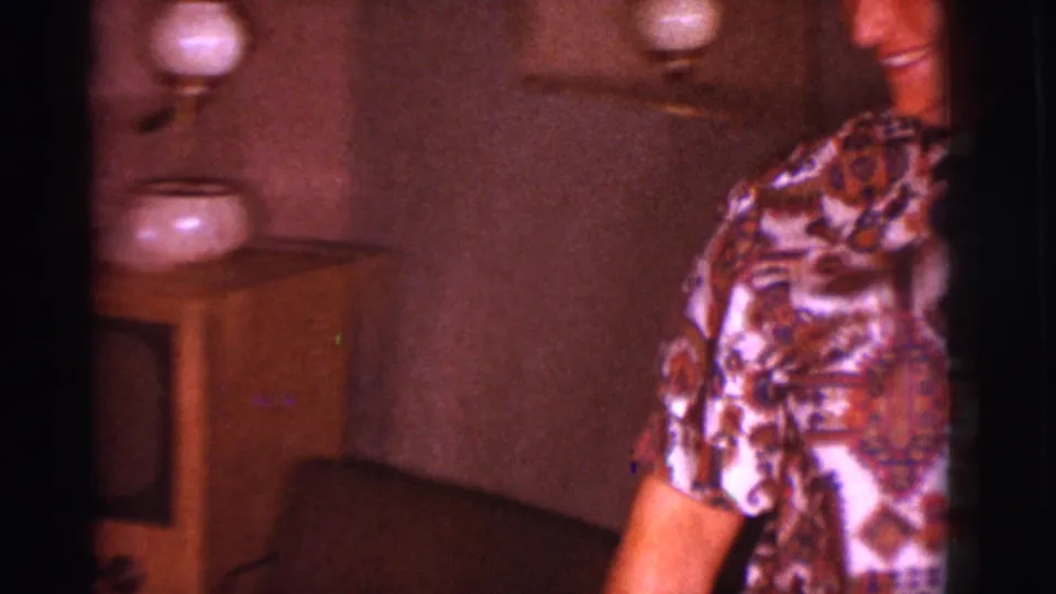 A person in a patterned blouse is partly framed beside a small table and lamp in the same 1960s home interior.