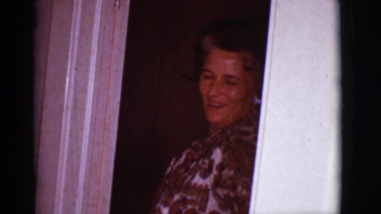 Woman in a patterned blouse smiling from a doorway inside a dim room.