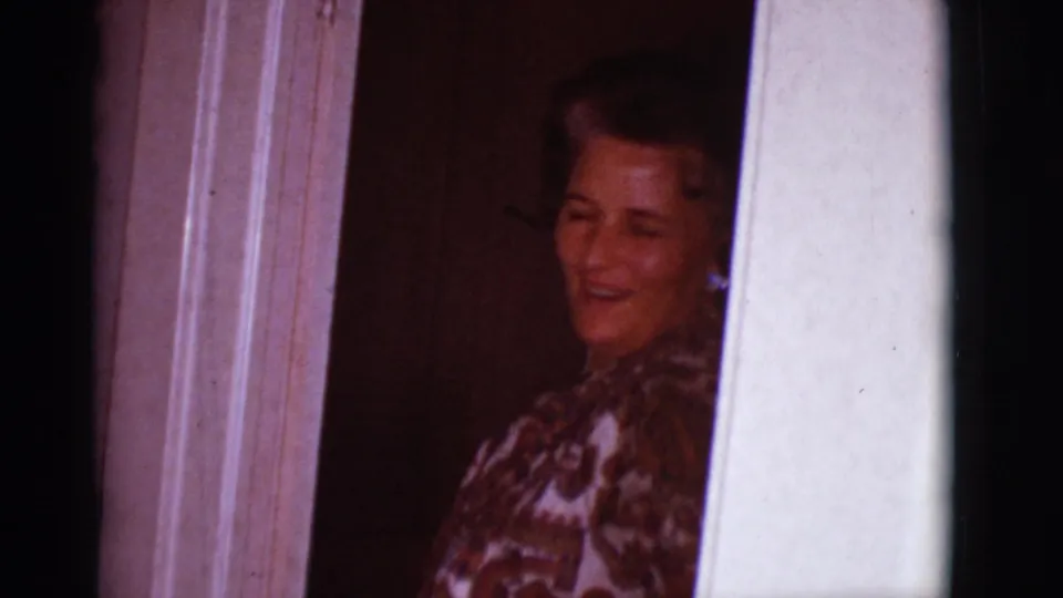 In a 1966 Clarkdale, Arizona home-movie frame, a woman smiles from behind white doorway trim in a dim interior.