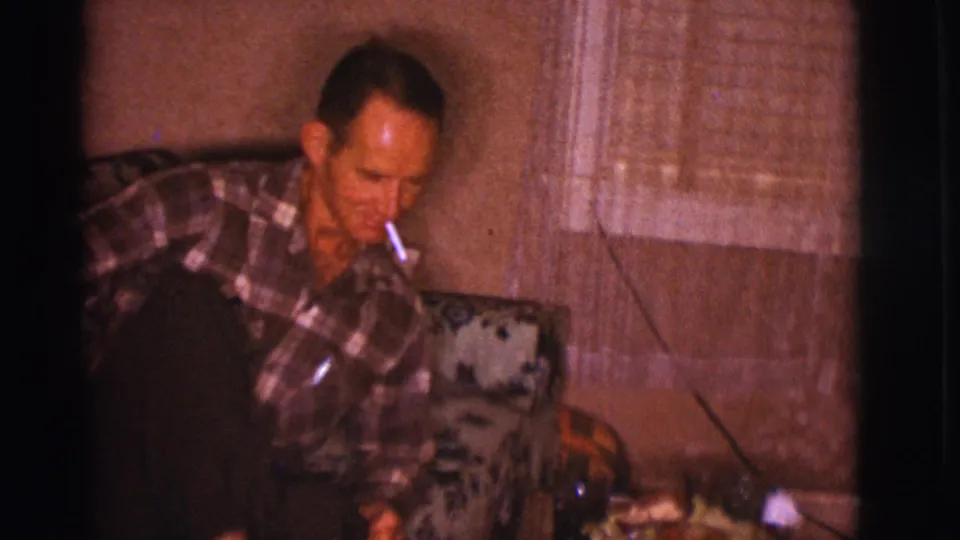 A man in a plaid shirt sits indoors on a floral sofa, smoking a cigarette in this 1966 Clarkdale home-movie frame.