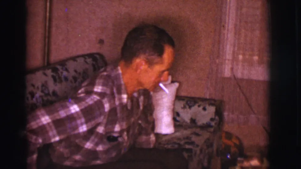 The man in a plaid shirt leans forward on a floral sofa, with curtains and household objects visible behind him.