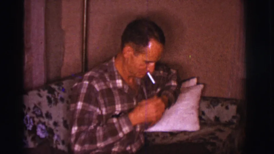 The man looks down and works with his hands, apparently preparing to light a cigarette in the 1966 home interior.