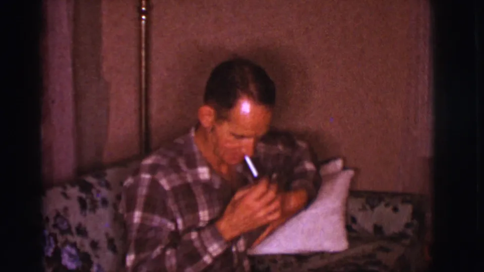A close home-movie view shows the man leaning forward on the floral couch with his hand near the cigarette in his mouth.