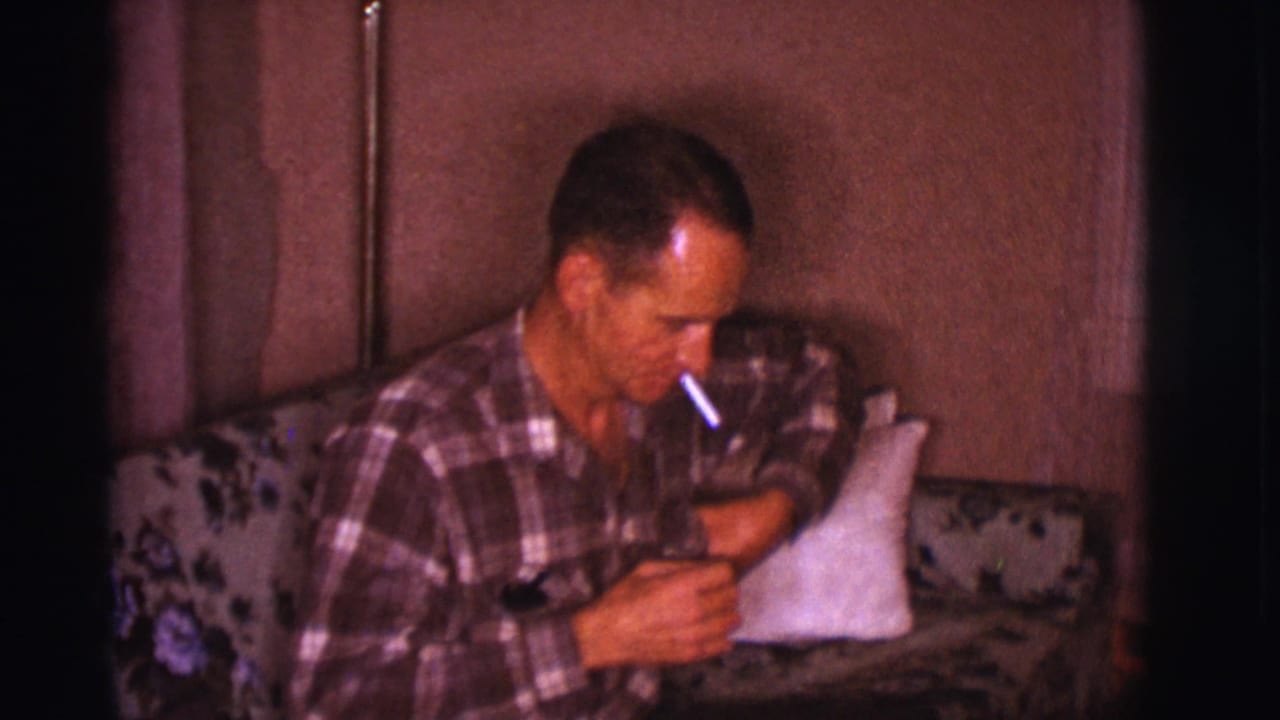 Man in plaid shirt seated on a floral couch with a cigarette.