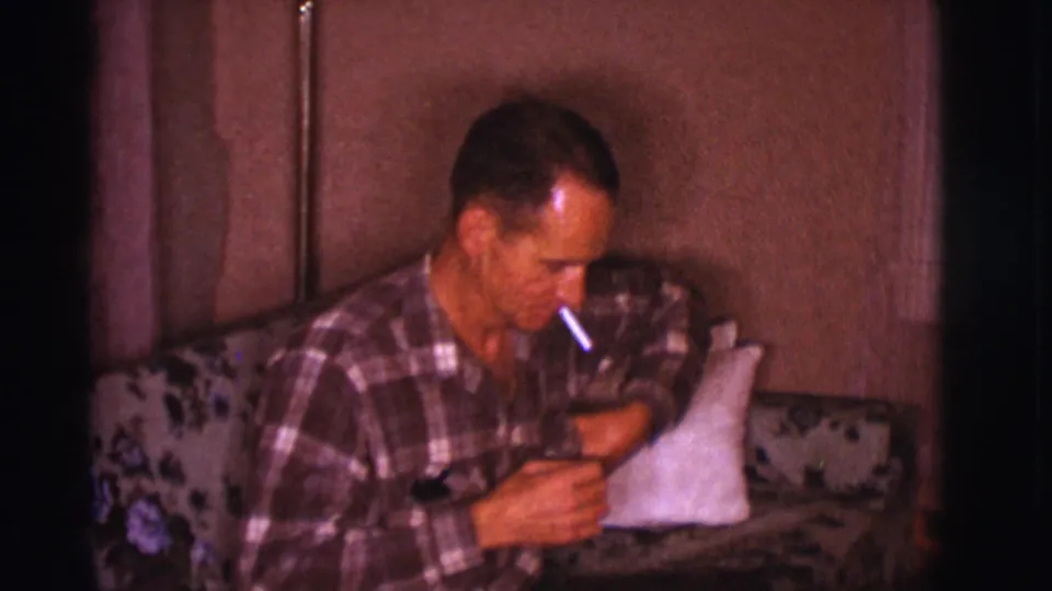In this 1966 Clarkdale, Arizona home-movie frame, a man in a plaid shirt sits on a floral couch holding a cigarette.