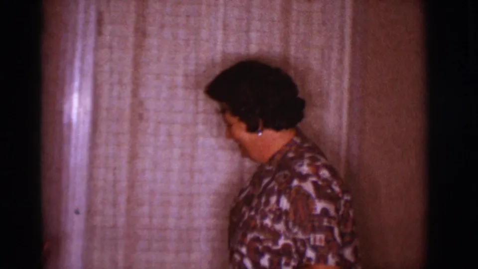 The woman turns toward a curtained window in the Clarkdale home, with soft 8mm grain and warm indoor color.
