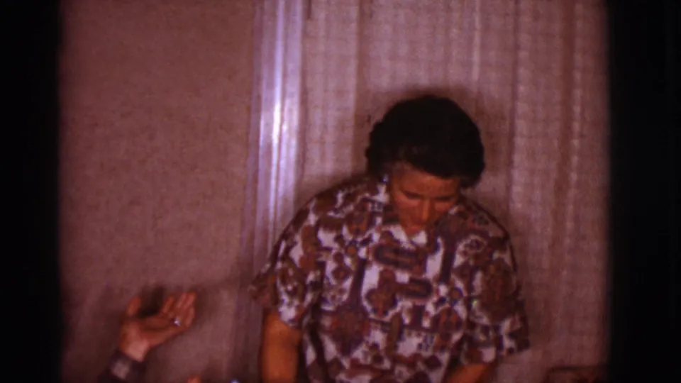 A woman in a patterned blouse looks downward near curtains while another person's hand enters the Clarkdale home-movie frame.