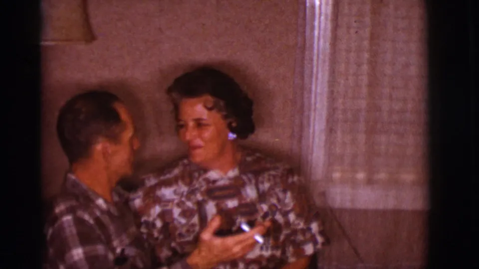 A husband and wife appear to smile and talk on a couch in a 1960s Clarkdale home interior, with curtains and a lamp nearby.