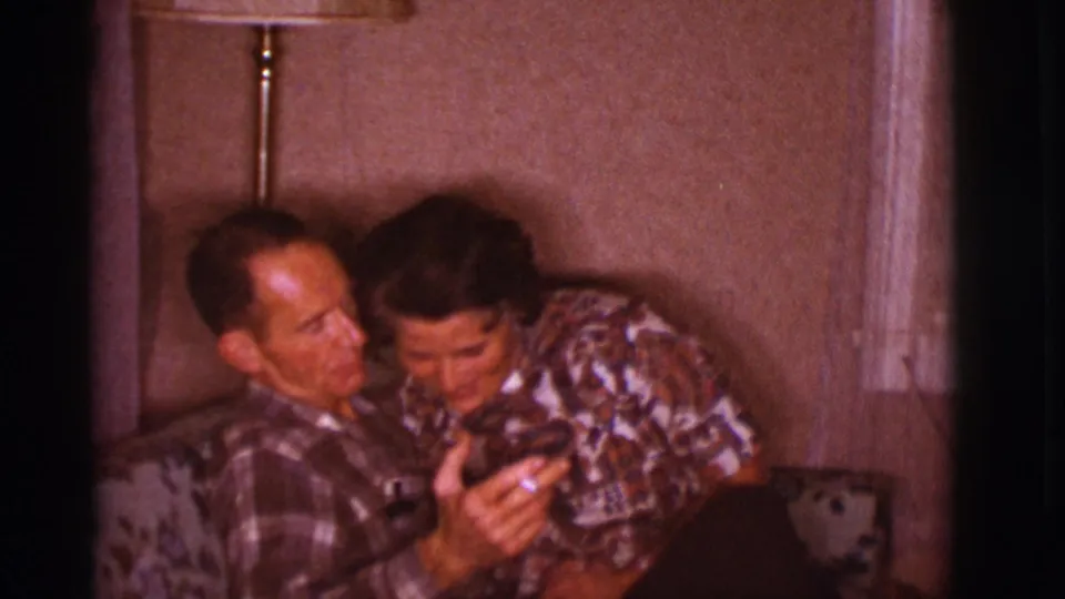 The man holds a cigarette while the woman leans into him laughing on a couch, captured as 1966 home-movie footage in Clarkdale, Arizona.