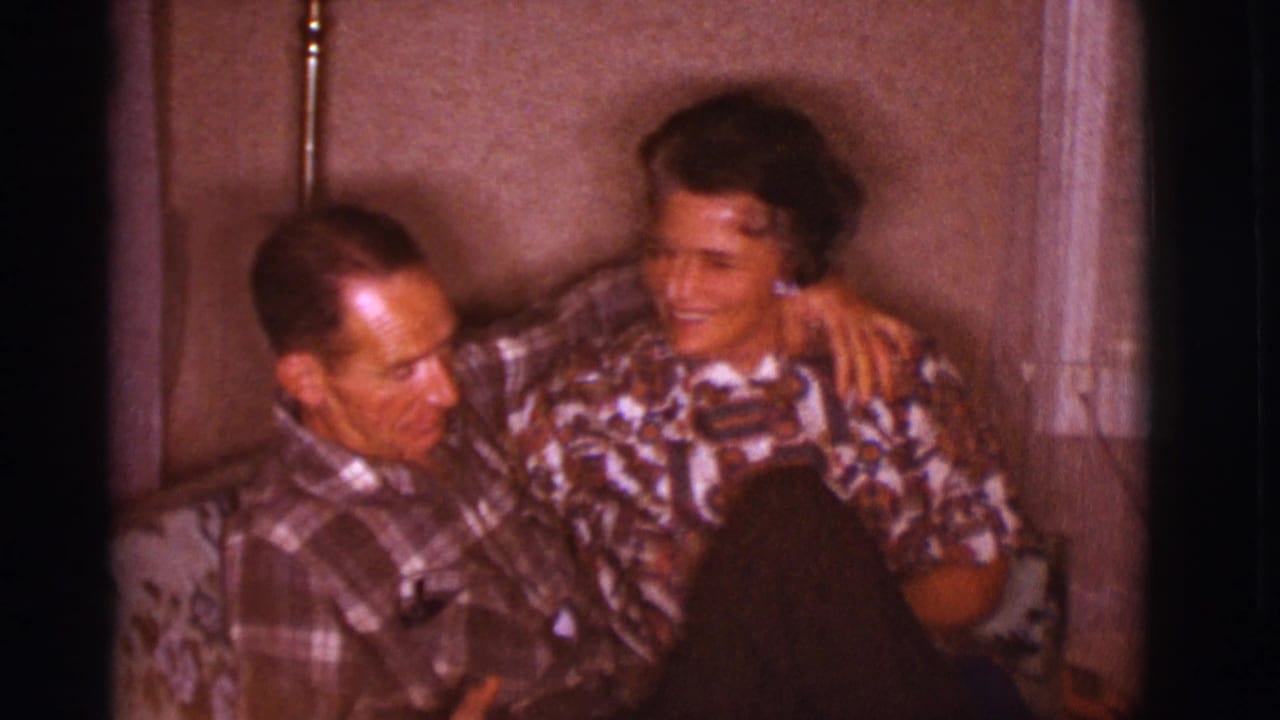 Two adults sit close together on a floral couch in a warmly lit room.