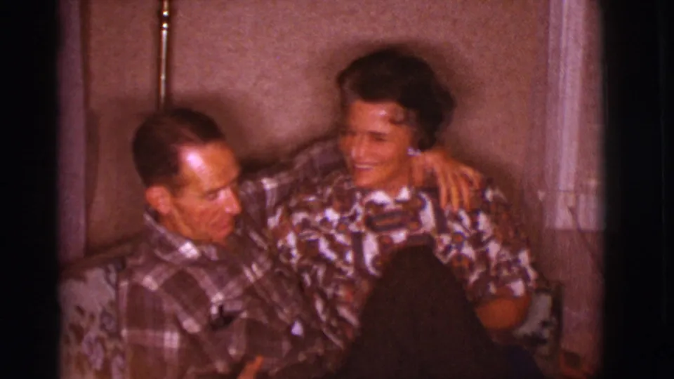 In a 1966 Clarkdale, Arizona home-movie frame, a man in plaid and a woman in a patterned blouse sit closely on a floral couch.