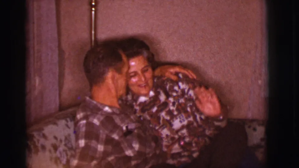 The man turns toward the woman as they sit closely on the floral couch, captured in soft 8mm color inside a private room.