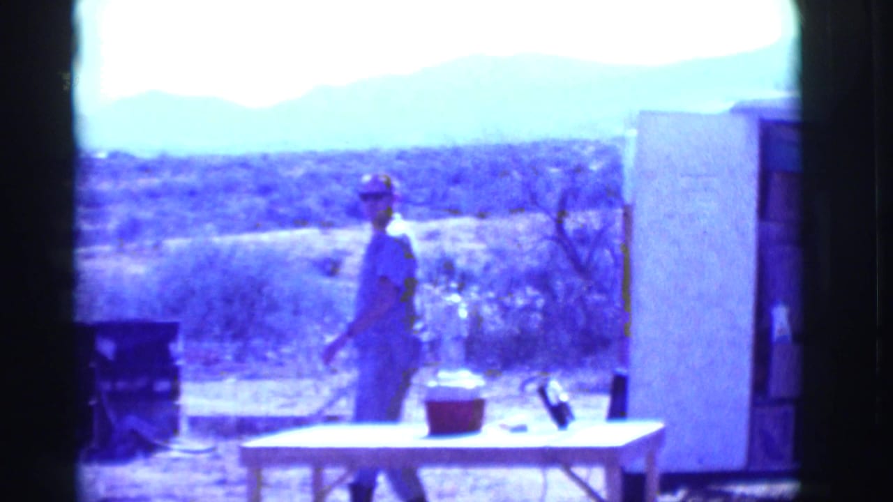 Man wearing sunglasses stands outside near a table and cooler in a desert yard.