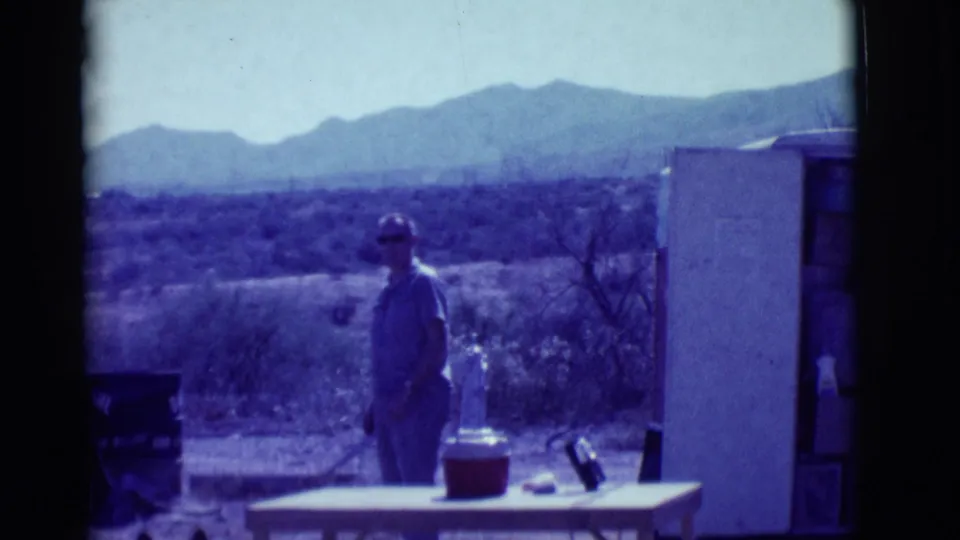 In a blue-shifted 8mm frame, a man stands in a desert yard near a table and cooler with mountains beyond.