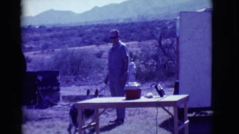 A man stands near a table and cooler in a Clarkdale desert yard as a small dog moves nearby, with mountains behind.