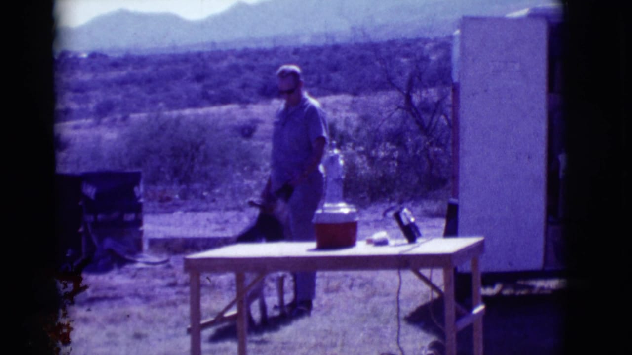 Man in sunglasses stands beside a German Shepherd near a folding table in a desert setting.