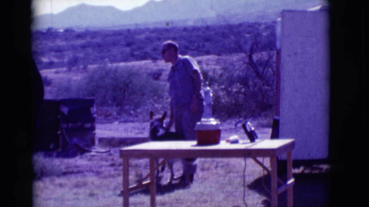 Man stands near a German Shepherd, folding table, cooler, and white trailer in desert scrub.