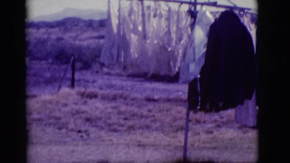 A clothesline with white laundry and dark garments stands in the Clarkdale desert, framed by dry ground, brush, and low mountains in 1966 8mm footage.