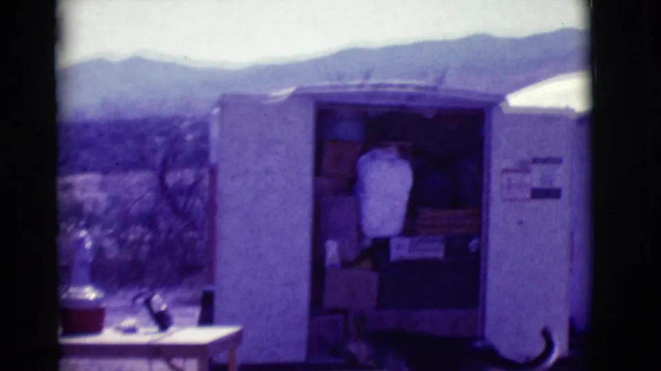 At the campsite, the German Shepherd stands near a small shed or trailer, with a table, supplies and distant hills visible.