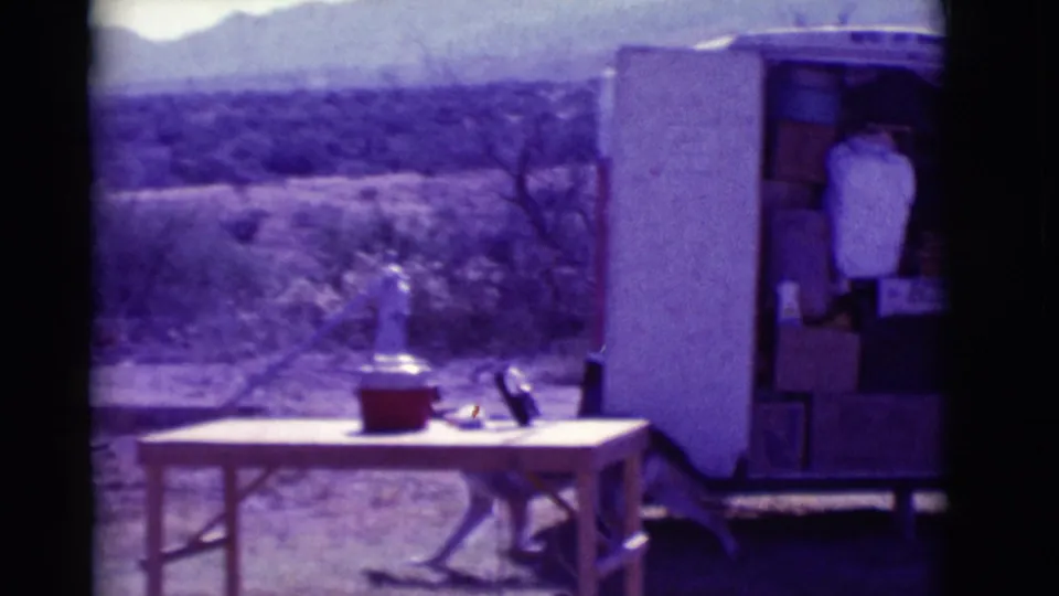 1966 Clarkdale, Arizona 8mm frame showing a table, red cooler, open trailer or storage area, desert scrub, and distant mountains.