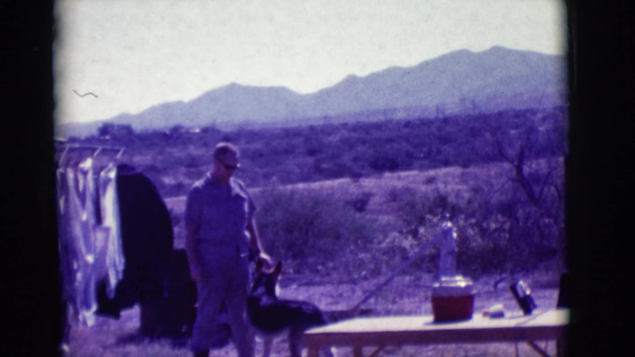 Man in light clothing stands with a dark dog near laundry and a table in the desert.